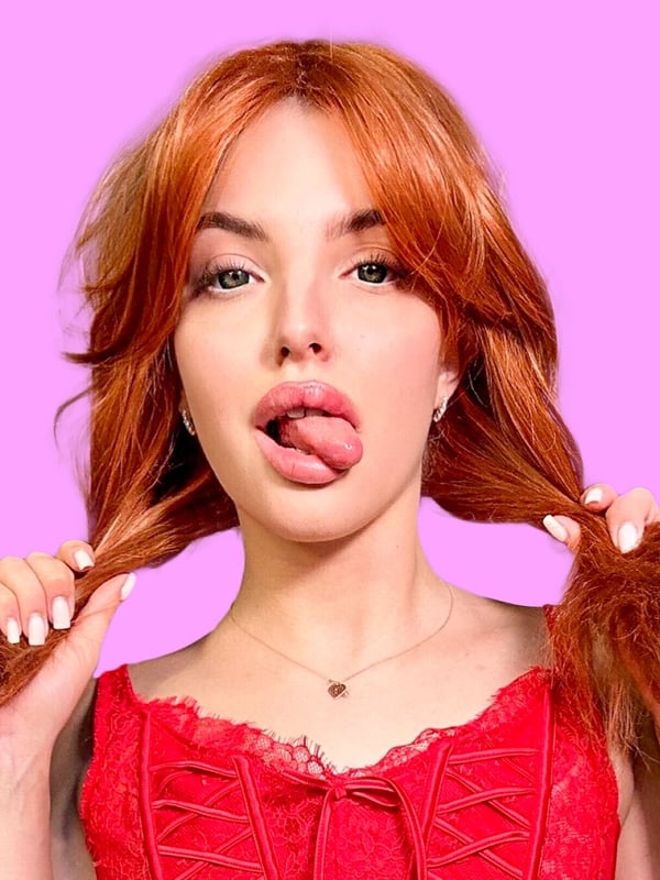 Close-up of a young woman with long red hair and green eyes, playfully sticking out her tongue while holding her hair, wearing a red top against a pink background.