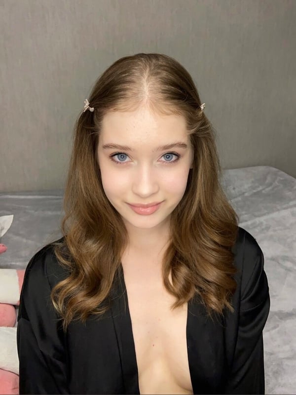 A young woman with wavy brown hair and hair clips, smiling softly while wearing a black robe on a bed.