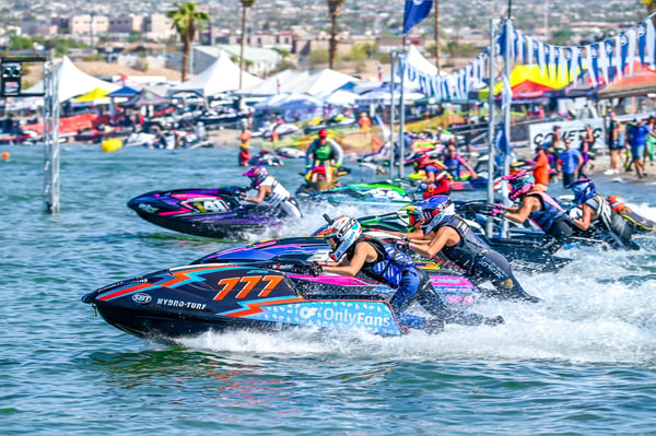 Dynamic scene of jet ski racers speeding across water during a competition event with colorful jetskis, spectators, and palm trees in the background.