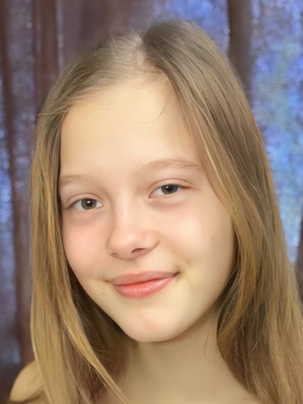 Young woman with long straight brown hair and a gentle smile, posing with a soft expression against a blurred curtained background.