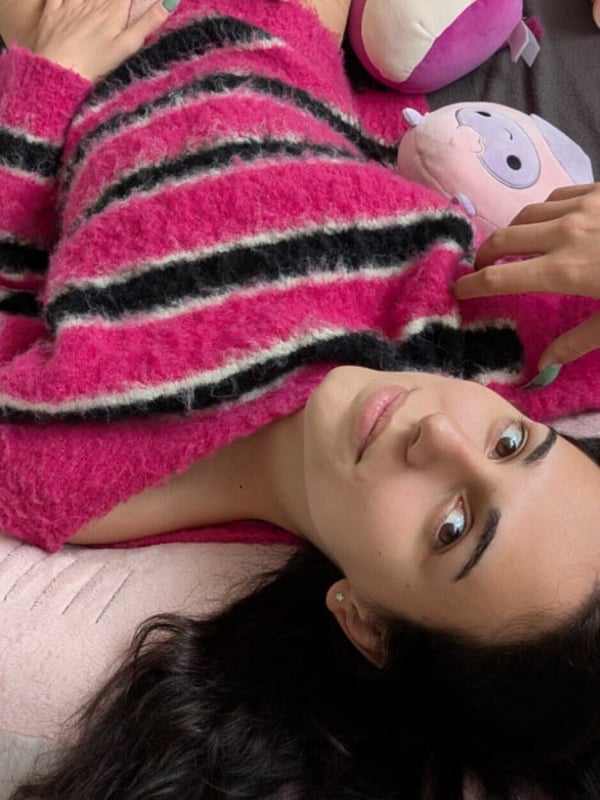 Close-up of a person with dark hair and striking eyes lying on a bed, wearing a striped pink and black fuzzy top, with plush toys nearby.