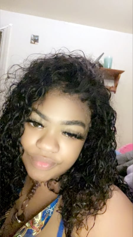 Young woman with long curly black hair smiling at the camera in a casual indoor setting with home decor in the background.