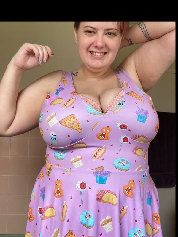 A smiling woman with short blonde hair raising her arm while wearing a vibrant purple dress with food-themed prints, standing indoors.