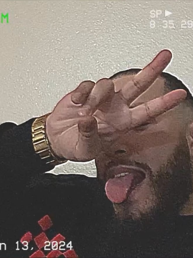 A man with a beard making a hand gesture and sticking out his tongue while wearing a black top and gold watch in a casual indoor setting.