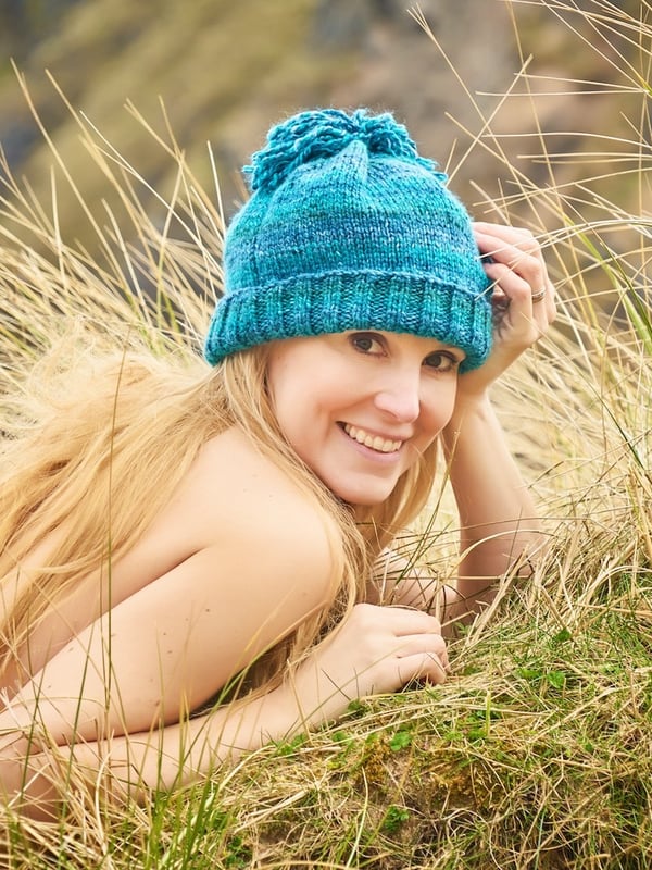 A smiling blonde woman wearing a blue knit hat, lying on tall grass in a natural outdoor setting.