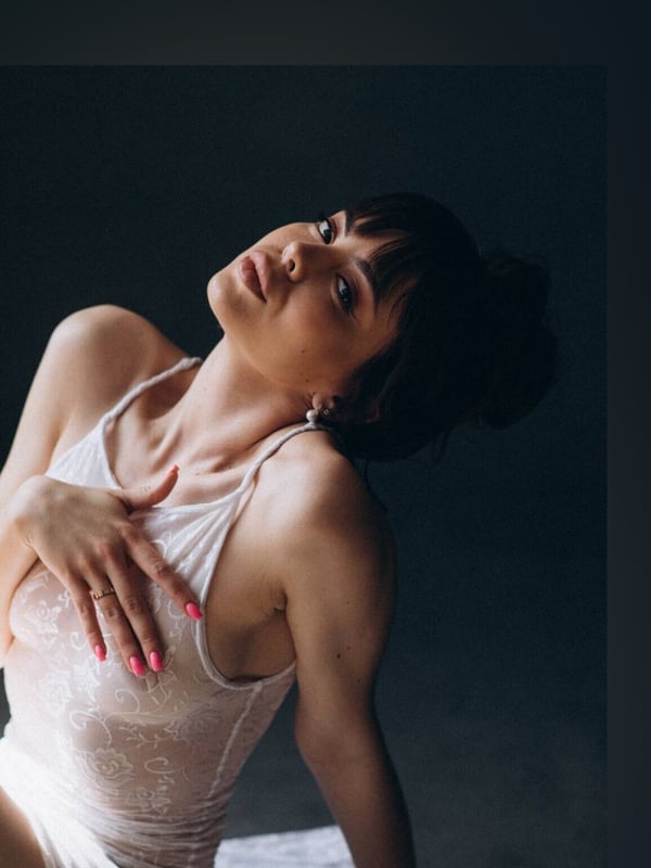 A woman with dark hair in a bun, wearing a white lace top, tilting her head back with a subtle expression in a dimly lit setting.