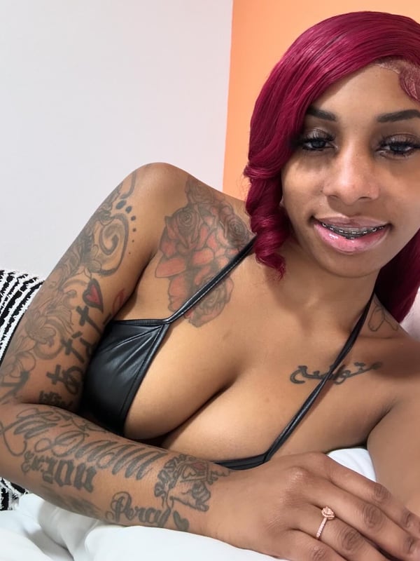 A woman with long red hair and tattoos posing confidently on a bed against an orange wall.