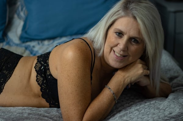 A woman with blonde hair reclining on a bed with blue pillows, wearing black lace lingerie and smiling at the camera.