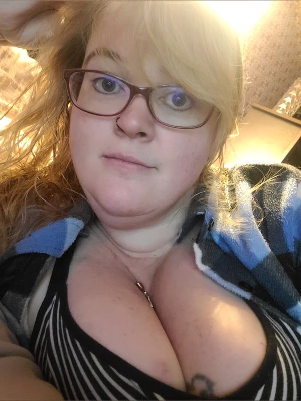 Woman with blonde hair and glasses, wearing a plaid shirt over a striped top, posing indoors.