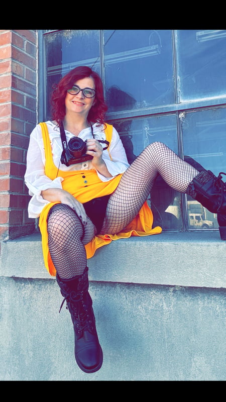A woman with red hair and glasses posing playfully on a ledge outside a building, wearing a yellow overall dress, white shirt, fishnet stockings, and boots while holding a camera.
