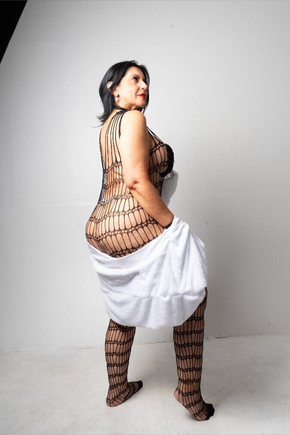A woman standing in a patterned outfit with a white cloth draped over, posing confidently in front of a plain background.
