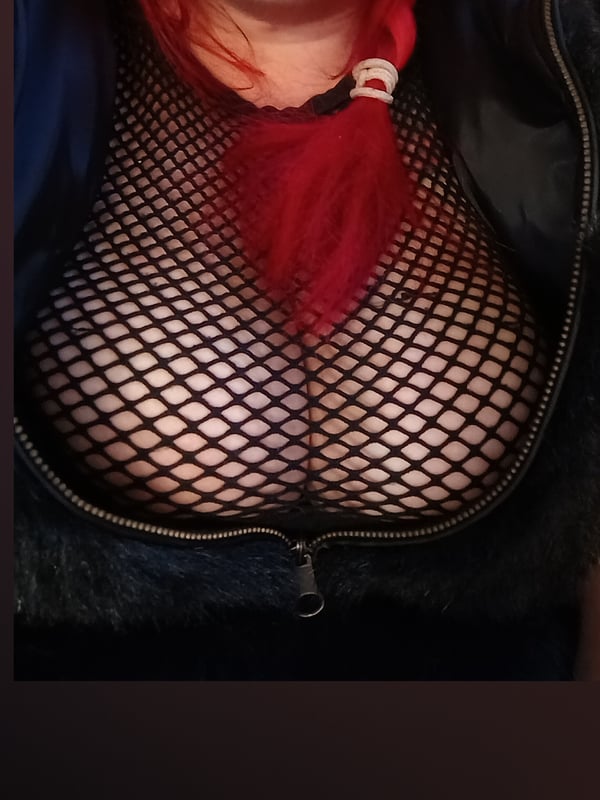 Woman with vibrant red hair wearing a sheer mesh top and black jacket, posing confidently with a zipper detail.