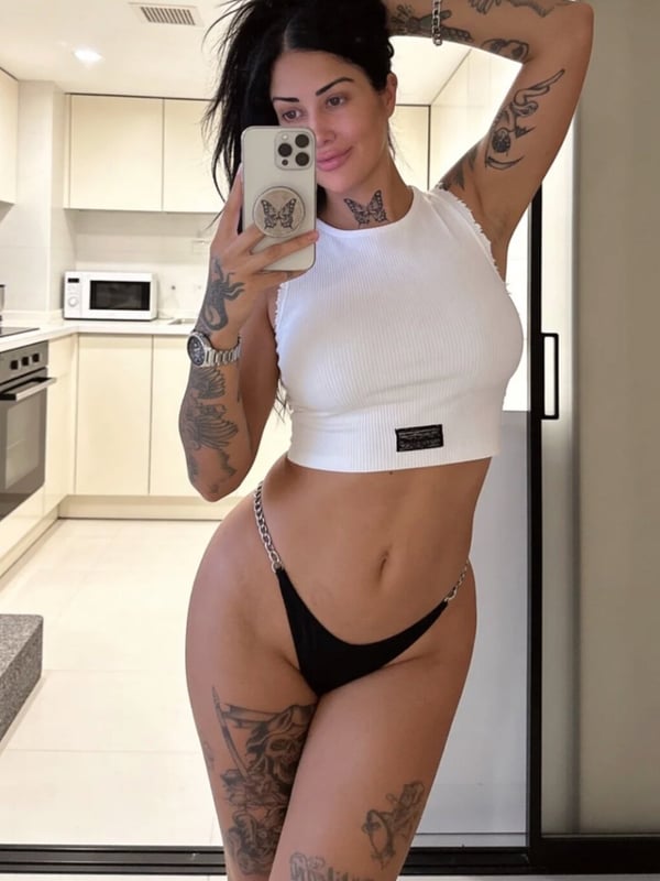 A woman taking a mirror selfie in a kitchen, wearing a white crop top and black bottoms, showcasing tattoos on her arms and legs.