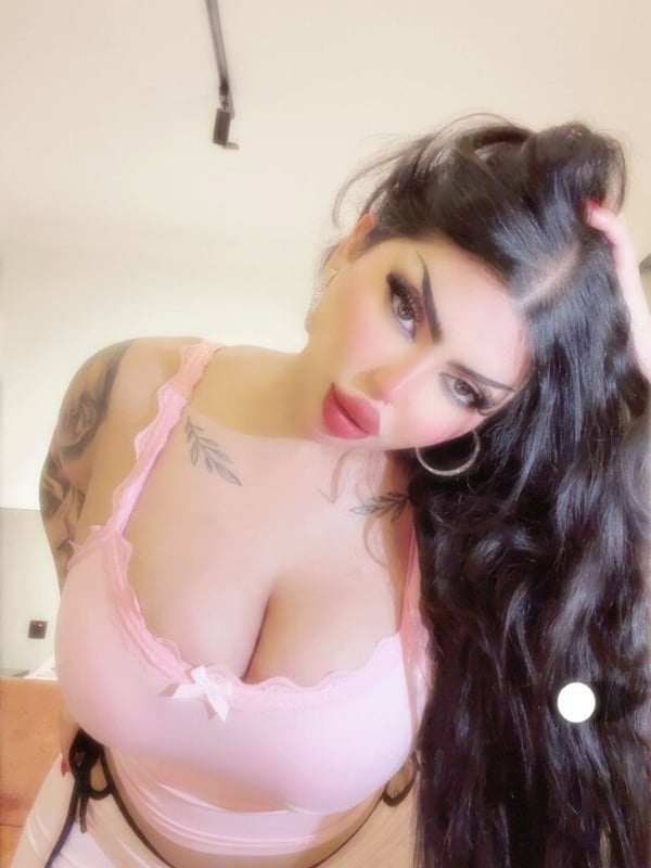 A woman standing in a room wearing a pink top and pants, with long dark hair and bold makeup.