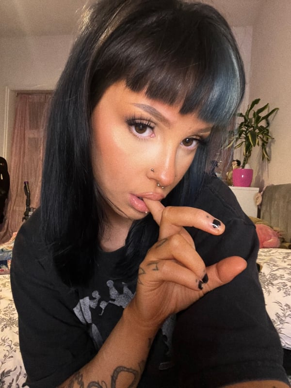 Young woman with long dark hair featuring blue highlights and bangs, wearing a black t-shirt, posing playfully with her finger to her lips in a bedroom setting.