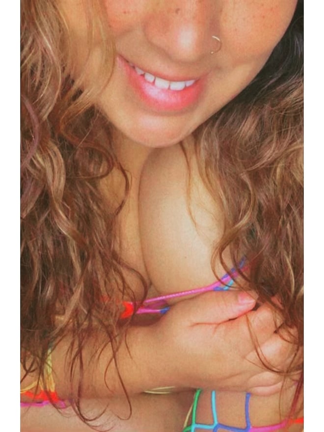 Close-up of a smiling young woman with wavy red hair, freckles, a nose piercing, and colorful bracelets on her wrists.