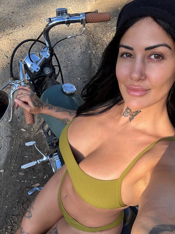 A woman wearing a beanie and bikini top posing next to a motorcycle in a dirt area.