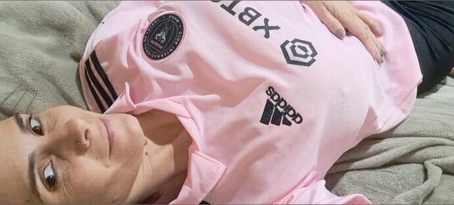 Woman lying on a bed wearing a pink Adidas polo shirt with logos, posing with hands on her stomach.
