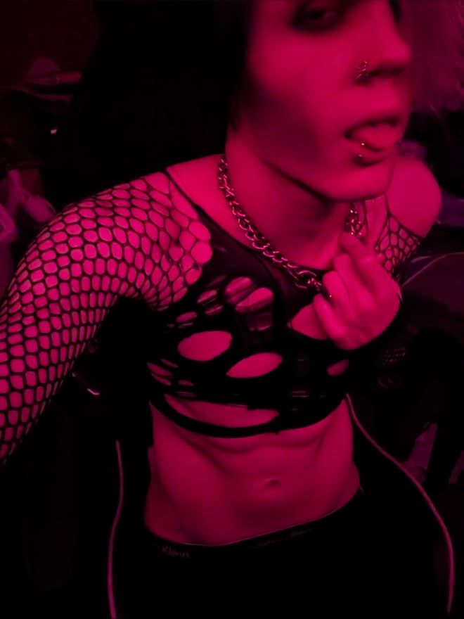 Person with heavy makeup and dyed hair posing in fishnet top and chains under pink lighting.