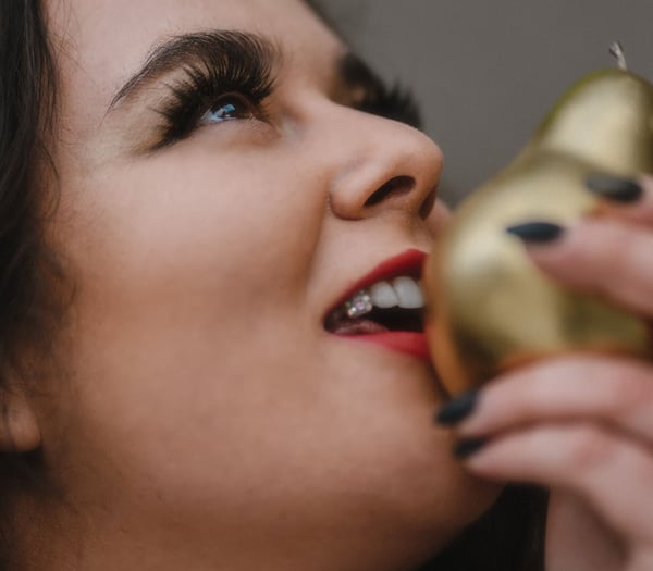 A woman with curly hair and bold makeup smiling while holding golden pear-shaped objects near her face.