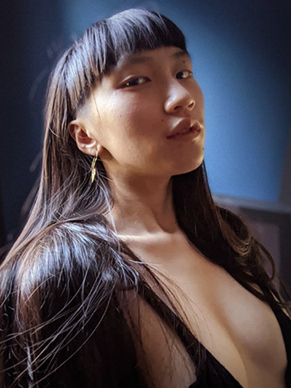 Close-up portrait of a young woman with long dark hair and bangs, wearing gold earrings, posing against a blue background.
