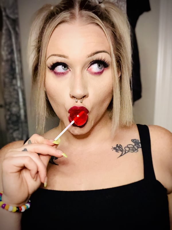 A young woman with blonde space buns and purple makeup holding a red lollipop to her lips, smiling playfully indoors.