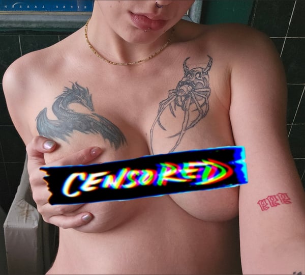 Young woman with extensive tattoos on her upper body posing topless in a bathroom setting with a censored overlay.