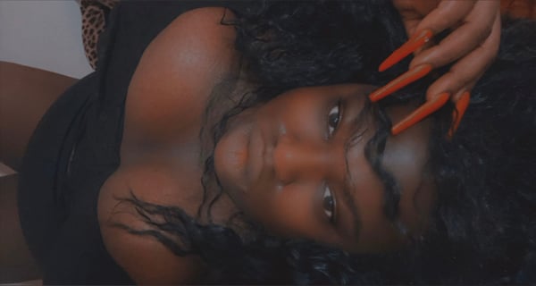 A woman with long dark hair and long nails posing sideways in a black top against a warm-toned background.
