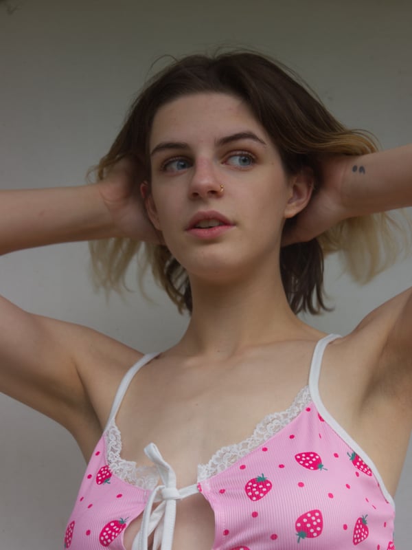 A young woman with shoulder-length blonde hair wearing a strawberry-patterned lingerie top, posing with hands behind her head against a plain background.