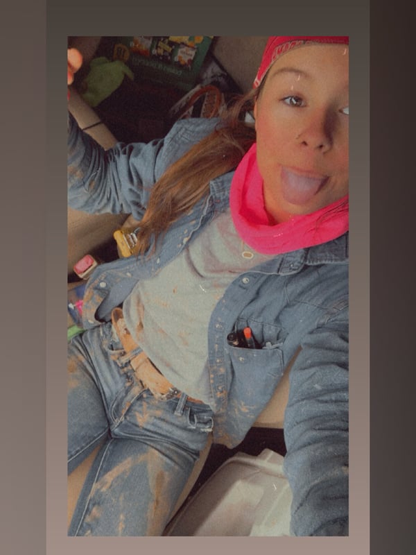 A young woman taking a selfie in a car, wearing casual denim clothing and a pink headscarf, smiling playfully.