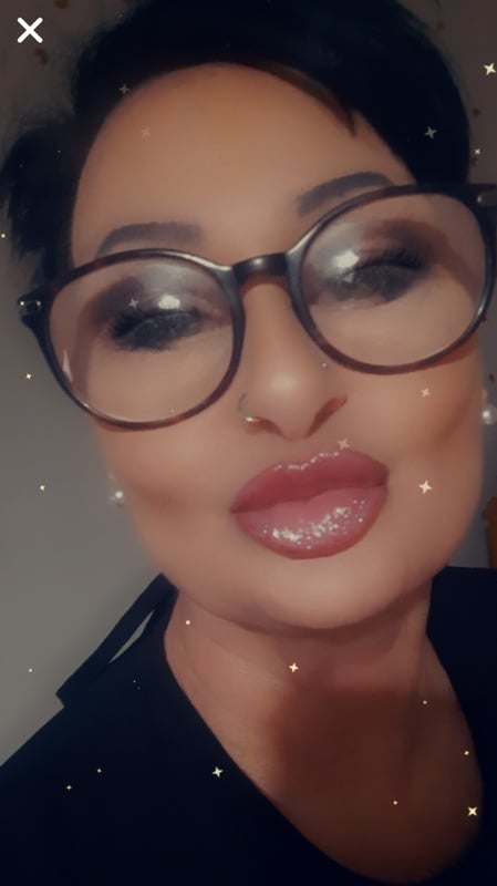 Woman with short dark hair wearing glasses, making a playful pout with glossy lips, in a close-up selfie with sparkly effects.