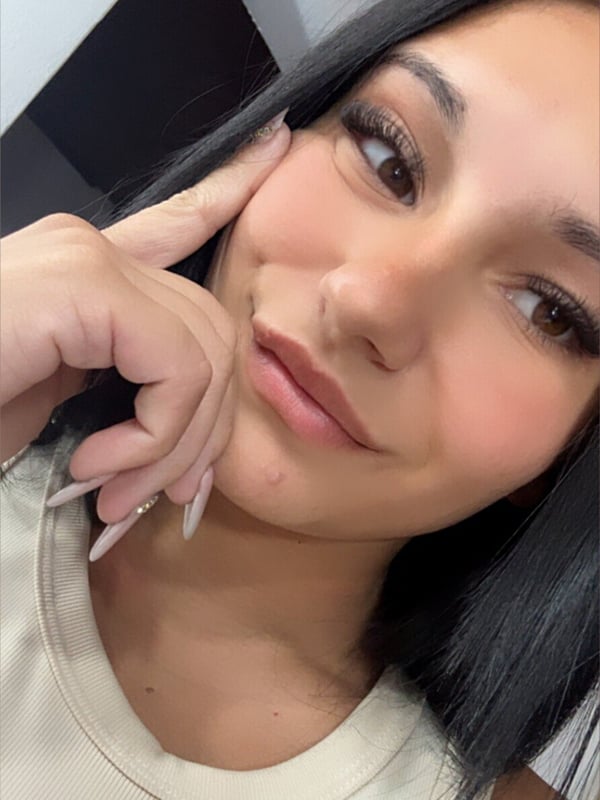 A close-up selfie of a young woman with long dark hair and manicured nails posing indoors.