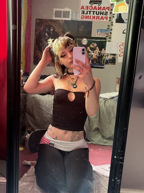 A young woman with split black and blonde hair making a peace sign, wearing a choker and open hoodie with tattoos on her chest, standing indoors with green lighting.