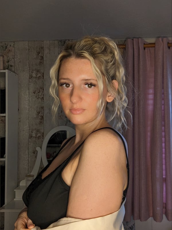 A woman with blonde hair in a ponytail stands in a bedroom wearing a black top and beige robe, smiling at the camera.