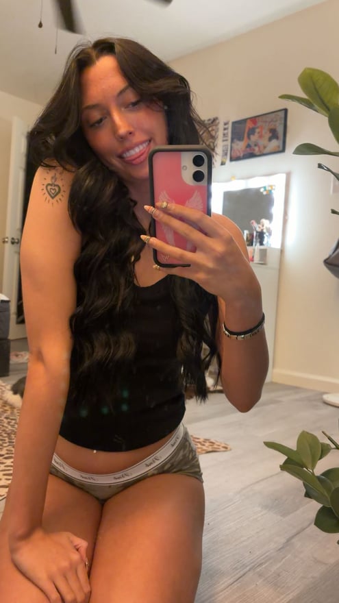 Young woman with long dark hair taking a mirror selfie in a bedroom, wearing a white top and gray shorts while sitting on a bed.