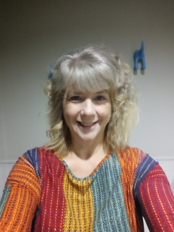 A smiling woman with curly blonde hair wearing a colorful striped top, posing indoors near a wall with a blue decorative item.