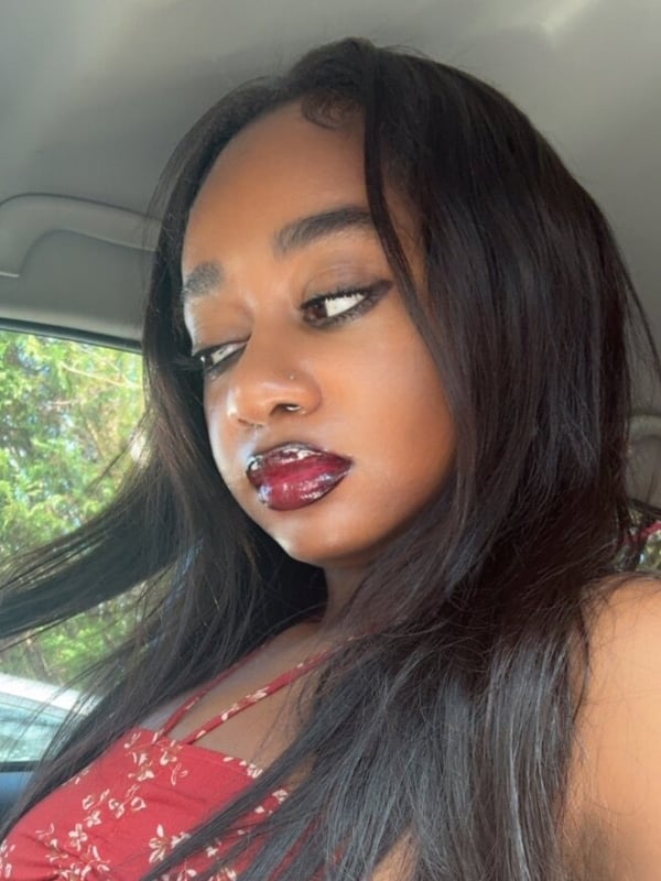Close-up portrait of a young woman with long black hair, red lipstick, and expressive eyes, posing in a car with a window view.