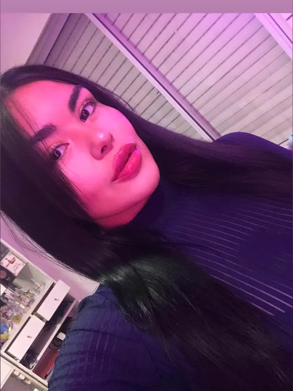 A young woman with long dark hair and bold makeup posing in a dimly lit room with purple lighting and a window in the background.