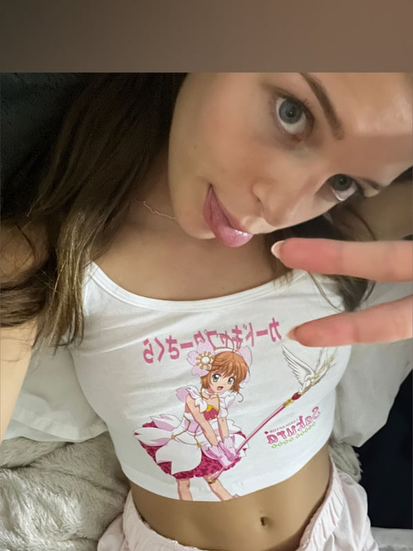 Young woman making a peace sign in a close-up selfie, wearing a graphic tank top and smiling at the camera.