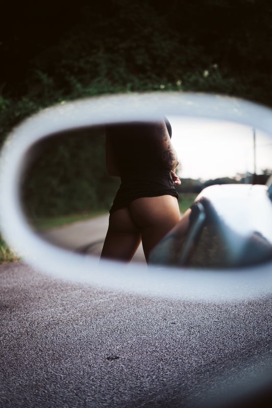 A person standing outdoors near a car mirror, reflected in the side view mirror against a green background.