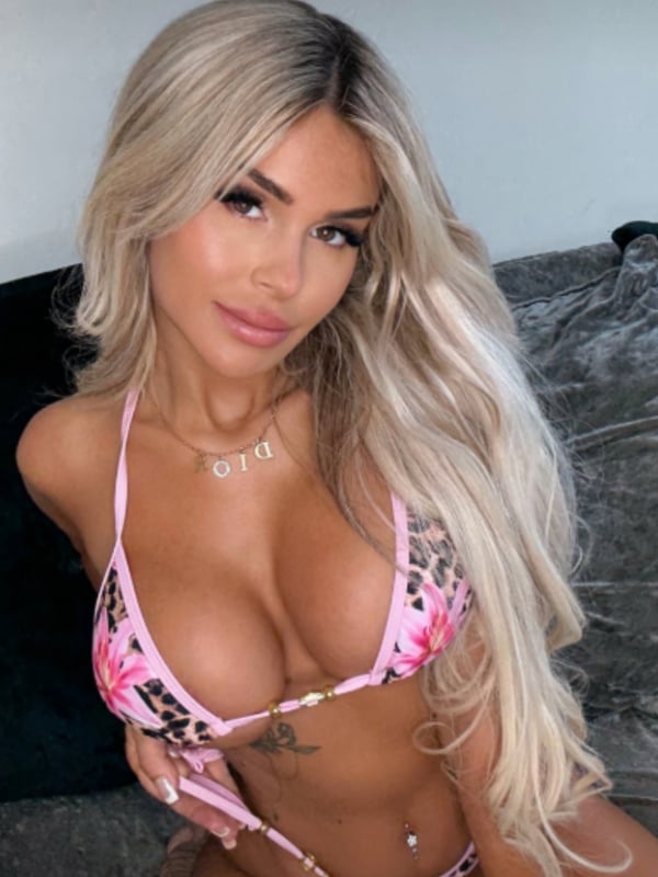 Young woman with long blonde hair posing confidently in a pink leopard-print bikini top, seated on a couch.