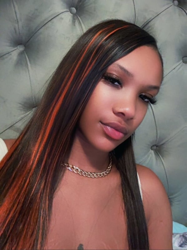 Close-up portrait of a young woman with long dark hair featuring orange highlights, wearing a gold chain necklace, posing against a gray tufted background.