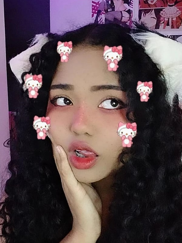 A young woman with curly hair and cat ear headbands in a pink-themed room with anime posters.
