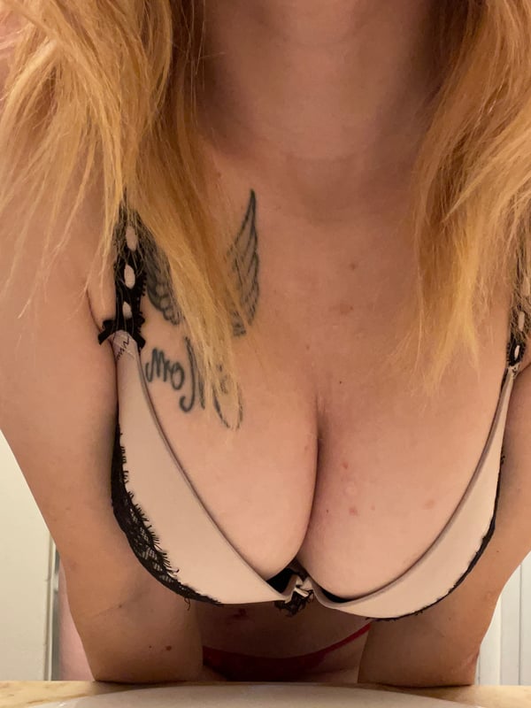 Close-up portrait of a blonde woman in a white lace bra, showcasing tattoos and a necklace.