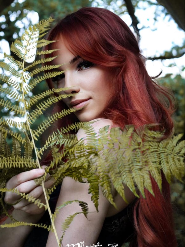 Young woman with long red hair smiling playfully while holding a large green fern leaf in front of her face, framed by a stone archway in a lush green forest setting.