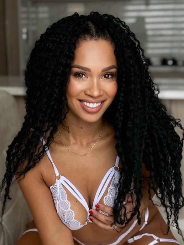 A smiling woman with voluminous curly black hair sitting in lingerie on a light-colored seat in a bright indoor setting.