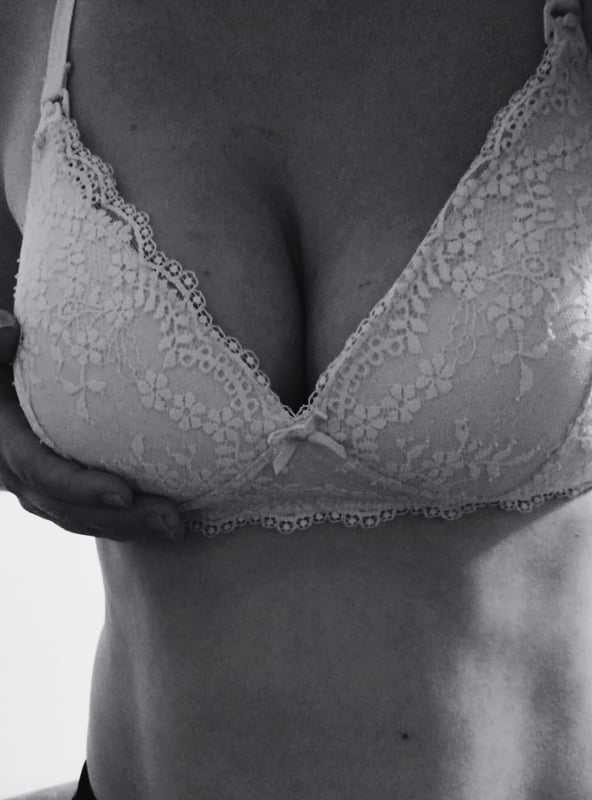 Close-up portrait of a woman in delicate lace lingerie, posing confidently with her hands on her chest against a light background.