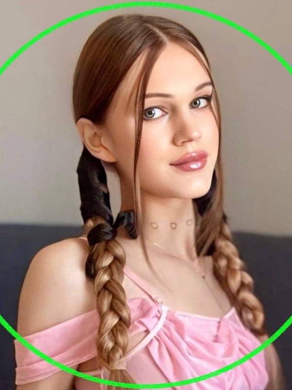 Close-up portrait of a young woman with brown braided pigtails, smiling softly, wearing an off-shoulder pink top against a dark background.