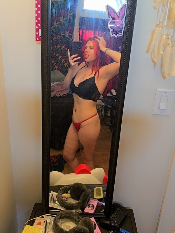Woman with red hair taking a mirror selfie in a black bra and red underwear, standing in a colorful bedroom with decorations and accessories.