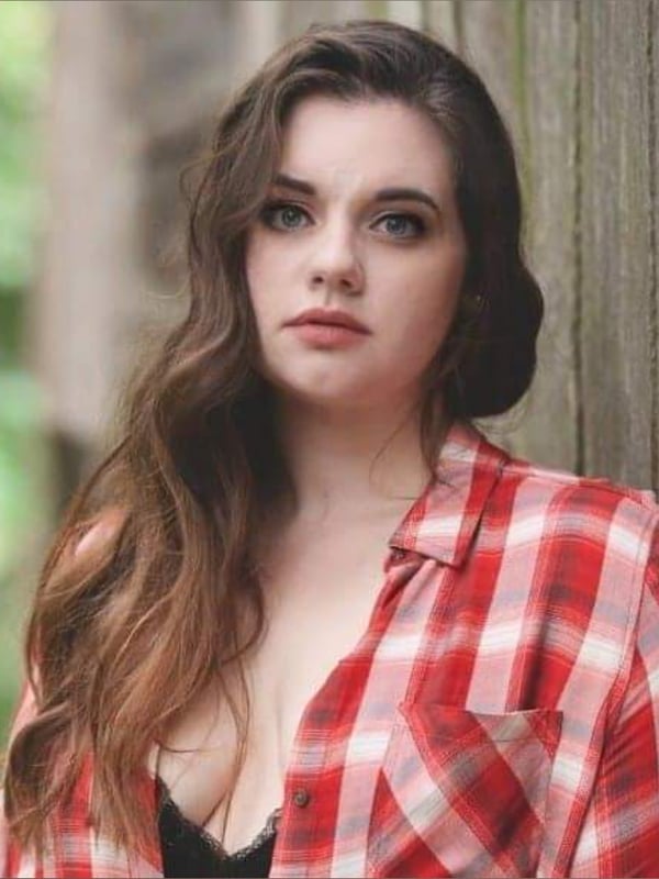 A woman with long wavy hair standing against a wooden wall in a plaid shirt, smiling softly in an outdoor natural background.